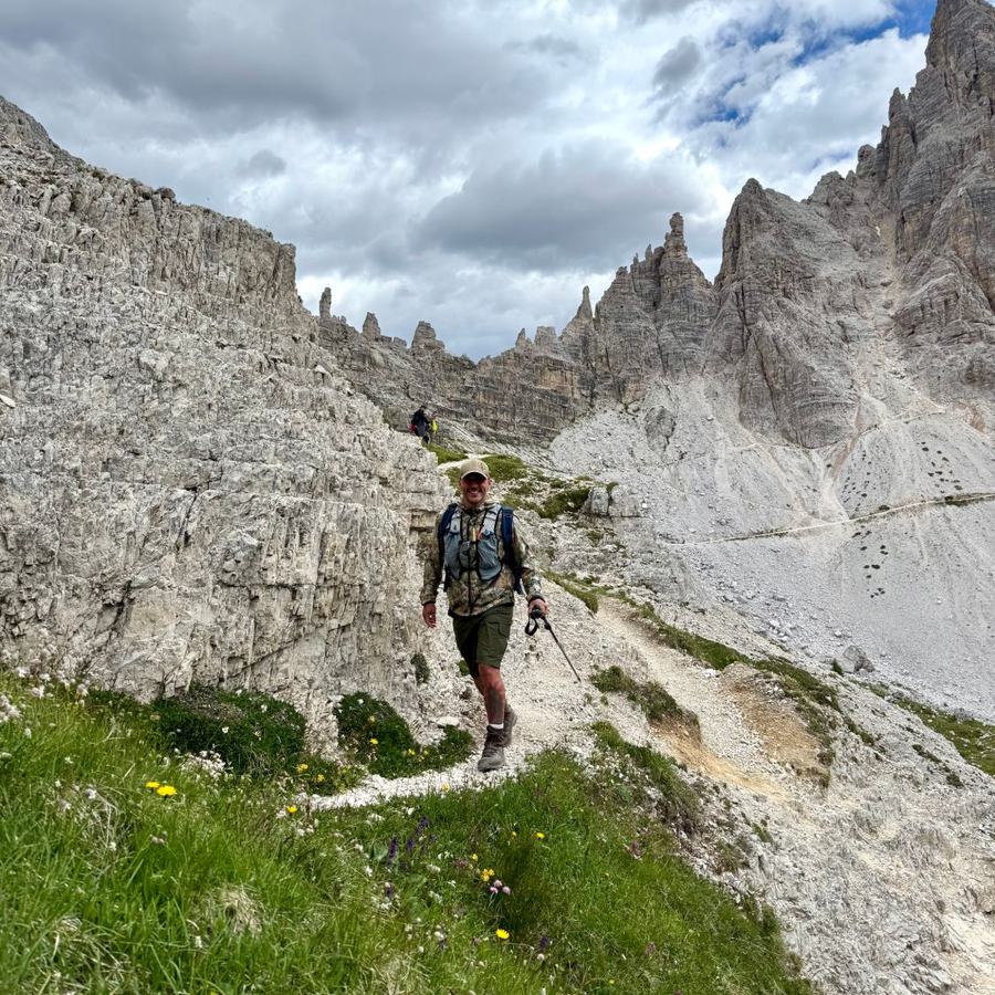 Scott Neve mid-stride on a mountain path below the limestone spires of the Dolomites, hiking pack on, grinning at the camera.